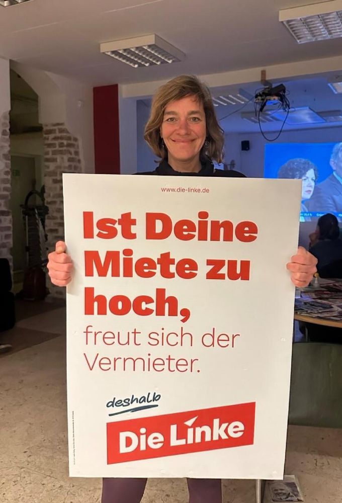 Jule hält ein Mietenplakat der Linken im linXXnet hoch und lacht fröhlich.