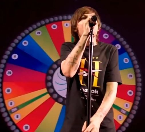 Louis Tomlinson (a pretty brunette man) singing in a mic wearing a t-shirt with a huge ass letter H and standing in front of a rainbow colored game wheel 