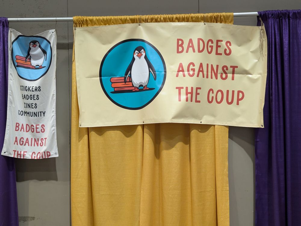 Two banners hanging on a wall.  One is portrait shaped and has a logo of a pissed off Penguin in front of a stack of books with "Badges Against the Coup.  Stickers. Badges. Zines. Community.". The other is larger and landscape shaped and has the same logo with the words Badges Against the Coup.