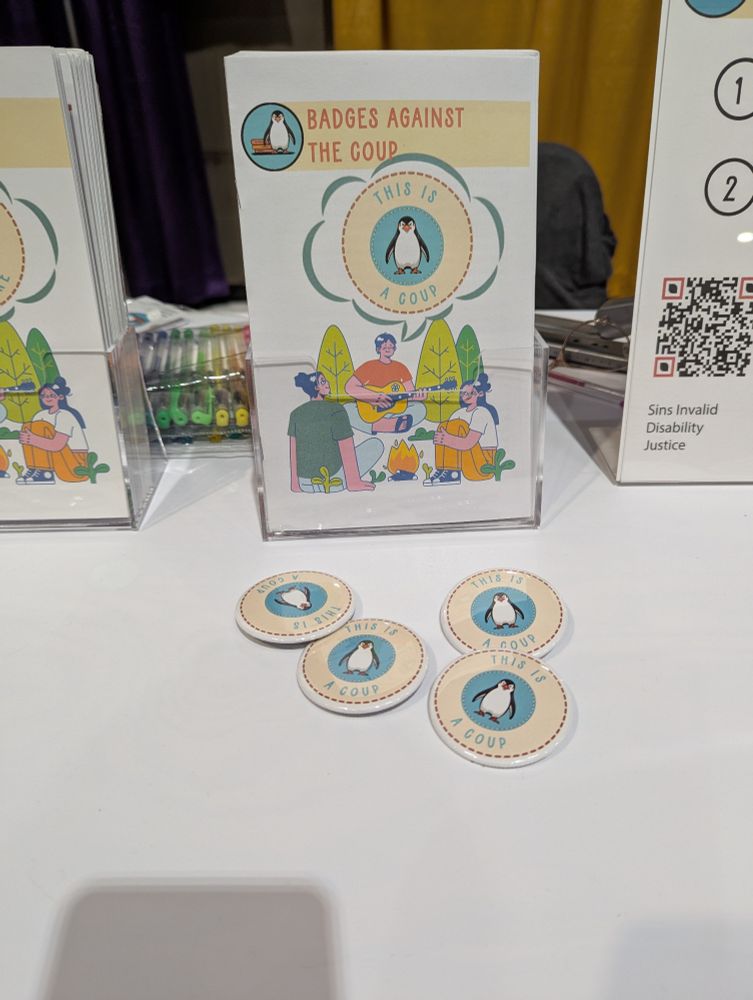 A zine on a conference table.  The cover has a group of young people sitting around a fire telling stories. At the top is an angry penguin before a stack of books with the words Badges Against the Coup.  The title of the Zone "This is a Coup" is in a speech bubble.  Under the zine is a four buttons with the penguin logo and the words This is a Coup.