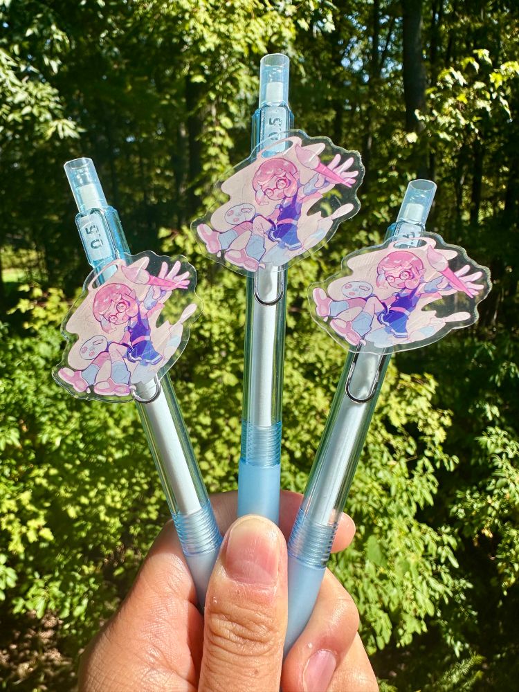 A hand is holding three blue pens which feature an acrylic design. The art shows an inkling wielding a color palette and brush. The backdrop is a forest scenery.