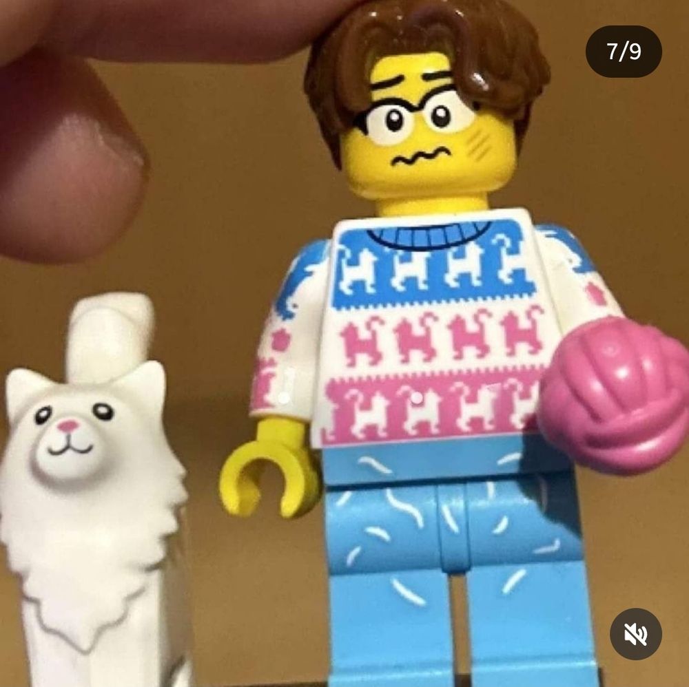 A photo of a LEGO minifigure, wearing notably a blue, white a pink striped jumper that resembles the transgender flag (with an additional cat motif), as well as seemingly gender-neutral face printing. Also has a pet cat and ball of yarn.