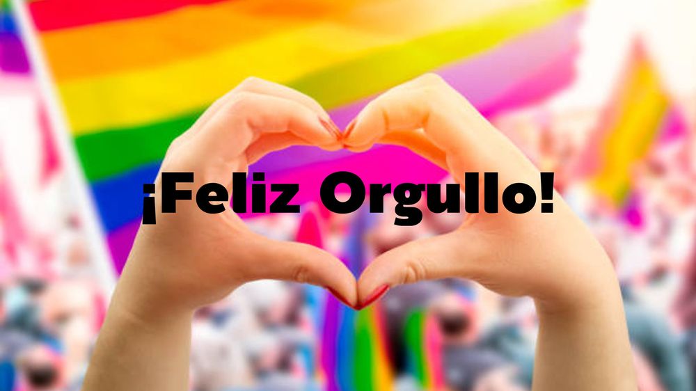 Manos formando un corazón. En el centro de la imagen, dos manos se unen para formar la silueta de un corazón. Al fondo, se ven banderas con los colores del arcoíris, típicas del movimiento LGBTQ+, y una multitud desenfocada. Sobre la imagen, en letras grandes y negras, está el texto: ¡Feliz Orgullo!