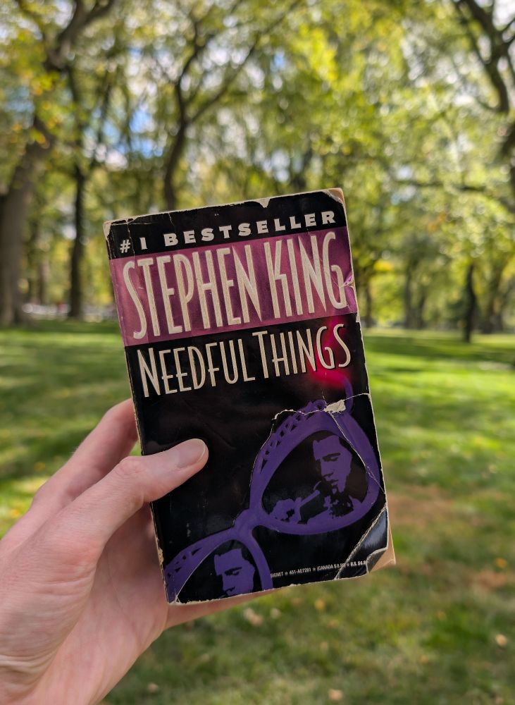 Book cover for Stephen King's novel Needful Things in front of green grass and trees.