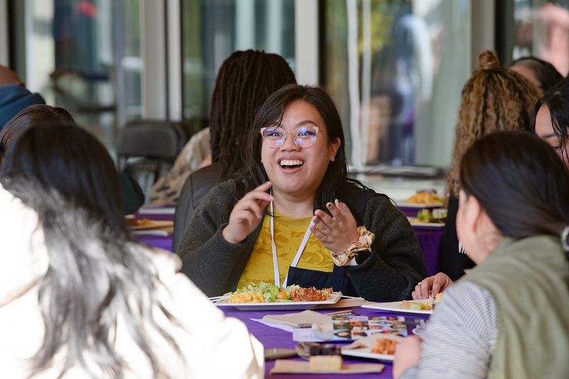 Smiling woman at an outdoor table with other women of color eating appealing lunch