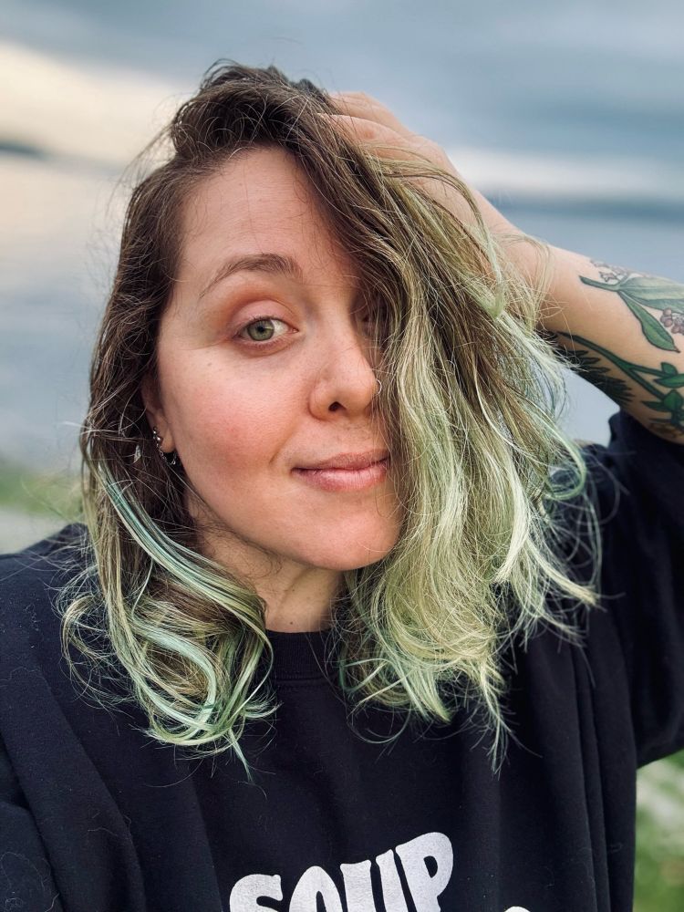A femme-bodied white person in an oversized black sweatshirt stands in front of the ocean. They have dark brown hair that fades into faded green, green eyes, and a sleepy smile on their face.