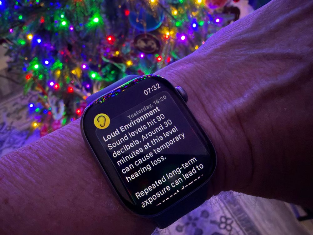 A person wearing a smartwatch holds up their wrist in front of a brightly lit Christmas tree with colorful lights. The Apple Watch screen displays a notification from the Noise app warning of a loud environment, noting that sound levels reached 90 decibels and that exposure at this level can cause temporary hearing loss.