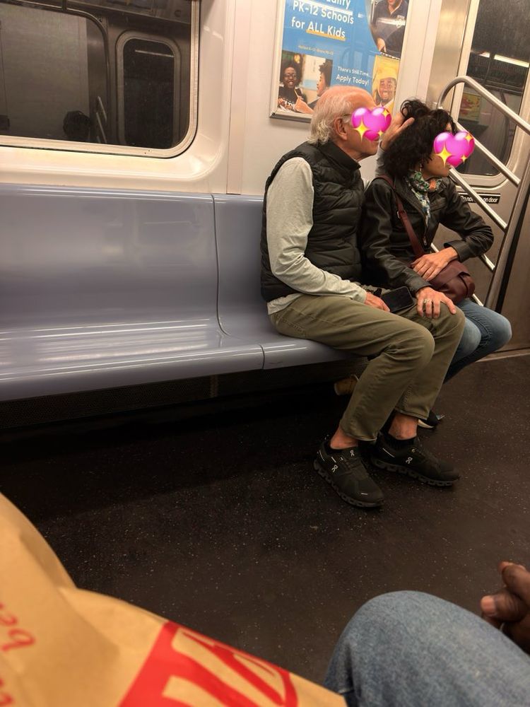 Two people sitting close together on a subway train seat, sharing an affectionate moment with their faces close together (faces obscured with heart emojis). They’re wearing casual clothing including black jackets and khaki/tan pants. The subway interior shows gray plastic seats, metal handrails, and an advertisement poster visible in the background. The photo is taken from across the aisle, with part of another passenger visible in the foreground. 
