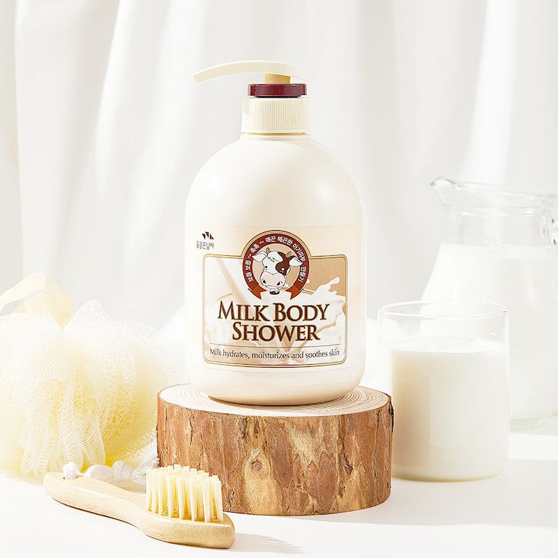White frosted bottle of Milk Body Shower with cow logo displayed on wooden pedestal with natural brush, yellow loofah, and glass of milk in background​​​​​​​​​​​​​​​​.