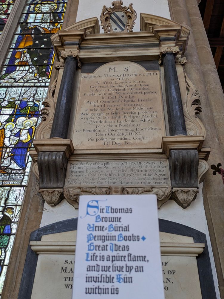 A wall mounted stone plaque on a church wall, commemorating the life of Sir Thomas Browne in Latin, and on a smaller plaque below, English.  Beneath both is held up a copy of his book on death and funerary rites, 'Urn Buriall', quoting from it the phrase 'Life is a pure flame and we live by an invisible Sun within us'.