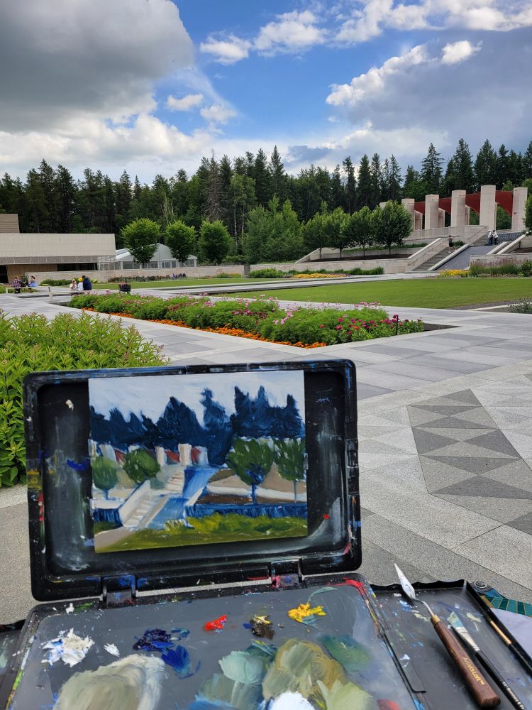 A painting sits on an easel with paint and a palette knife on the pallette. In the background is the Aga Khan exhibit at the University of Alberta Botanic Gardens