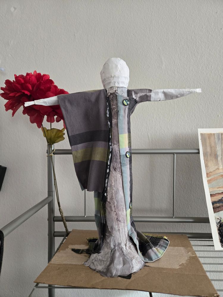 This is a vaguely human shape, standing on top of cardboard. I have chosen not to include legs, but the arms are visible near the top. The head is plain white for now. The fabric glued to the figure is plaid with the following colors: grey, green, and a hint of light blue. There are 3 matching buttons attached.