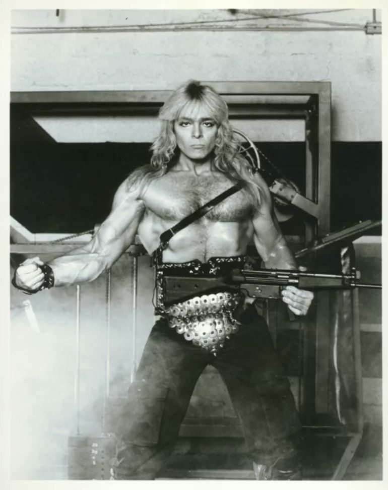 wieder ein schwarz weiß foto. hier ist er wieder mit nackter Bodybuilder Brust zu sehen. in einer Hand hält er ein  Maschinengewehr, mit dem andern Arm flext er