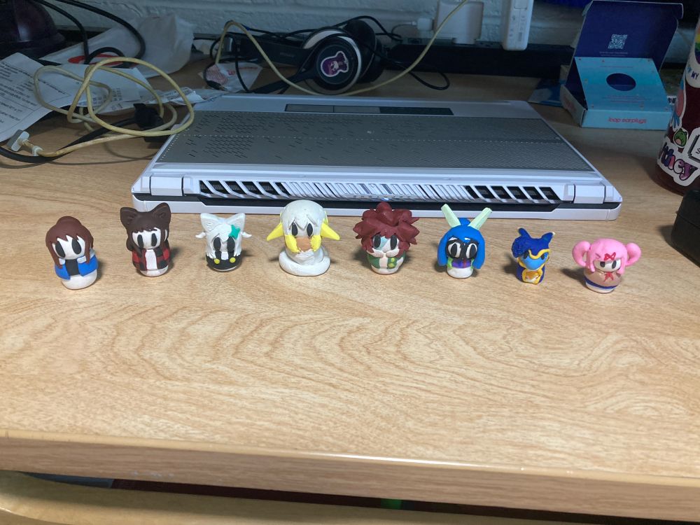a photo of a desk with eight small polymer clay figures. they all consist of a white cylinder body and a white sphere for a head, along with two black oval eyes and various other details. from left to right, they are: a figure with a brown ponytail, a black t-shirt, and a blue hoodie; a dark-haired catgirl with red flannel, a small white-haired foxgirl with a teal hairclip and black overall skirt; a snake-tailed girl with yellow pointed ears, hair that fades from white to yellow, and a beige hoodie; a zombie boy with a two-toned face, spiky red hair, and a patchy dark green jacket; a girl with a blue ponytail, round glasses, a lime green jacket, and a pair of robot ears sticking up from her hair; a dark blue avali with yellow highlights and a mass of feathery hair on the side of his head; and Natsuki from Doki Doki Literature Club, a pink-haired girl with pigtails wearing a beige school uniform and a blue skirt