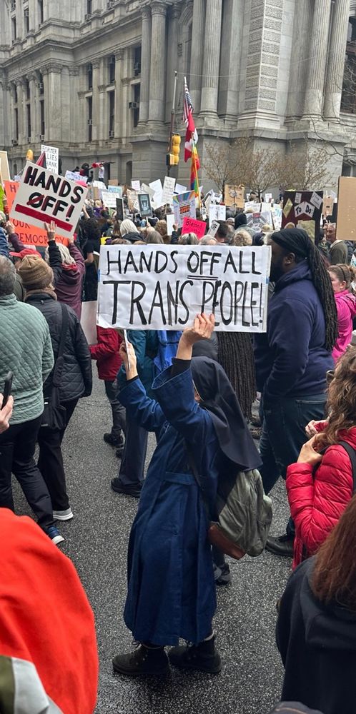 A protest in front of Philadelphia City Hall. Many people gathered, in the center a nun holds a handwritten sign that reads, HANDS OFF ALL TRANS PEOPLE!