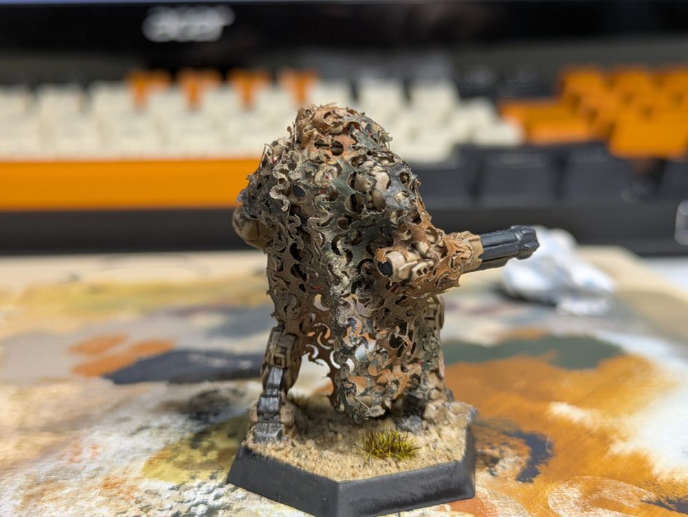 A desert camo Vindicator Battlemech with a red and white angular camouflage and a tattered camo net draped over its head and back (rear view)