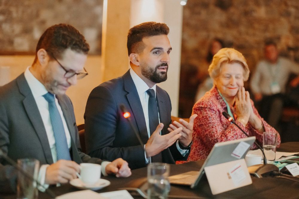 Three people are seated at a table, likely at a conference or meeting. 

The person on the left, a man, is wearing a dark gray suit and light blue tie. He is looking down, and seemingly engaged in the proceedings. He is holding a coffee cup and saucer in his hands.

The person in the middle, another man, is wearing a dark suit, and is speaking.  His hands are gesturing.

The person on the right, a woman, is wearing a reddish-pink, patterned jacket over what appears to be a light-colored top. She is smiling and holding her hands together in front of her face.

There are microphones on the table in front of each person and a light-colored tablet is present in front of the woman. The room appears to be a conference hall or similar meeting space with a light brownish-beige wall backdrop.  Blurred figures of other attendees are visible in the background.