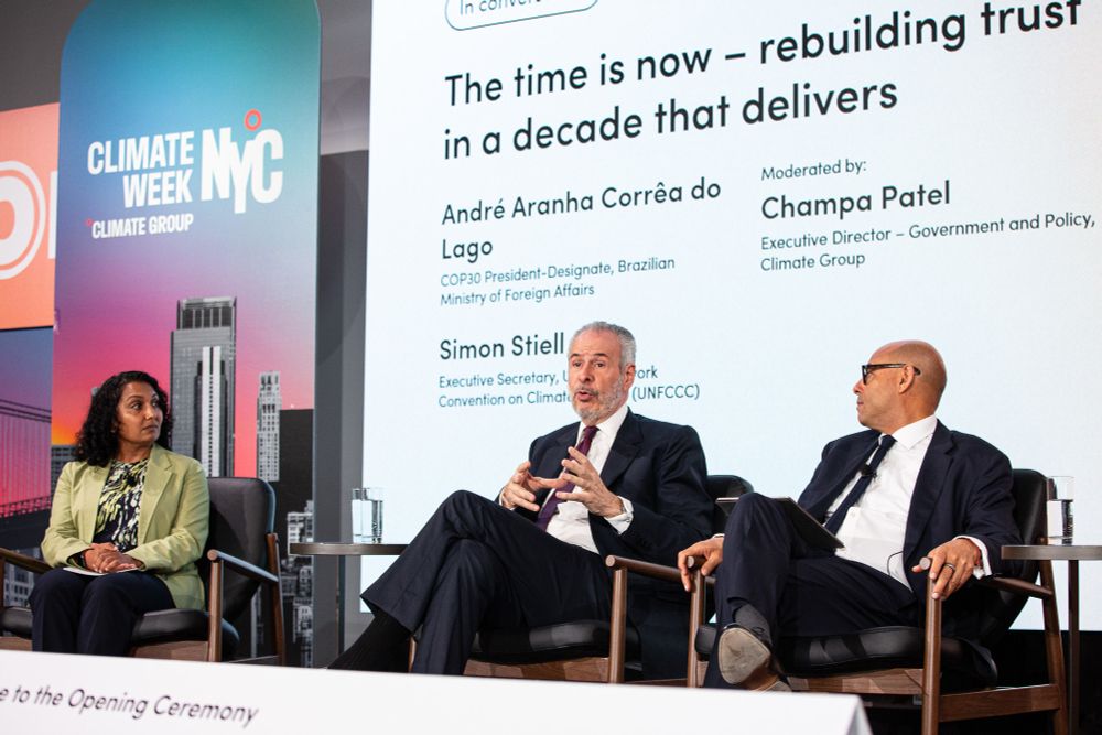 André Corrêa do Lago speaks on stage at Climate Week NYC, he is wearing a suit and tie and is joined by Simon Stiell, UNFCCC who is also wearing a suit and tie and Champa Patel from the Climate Group who is wearing a green blazer. 