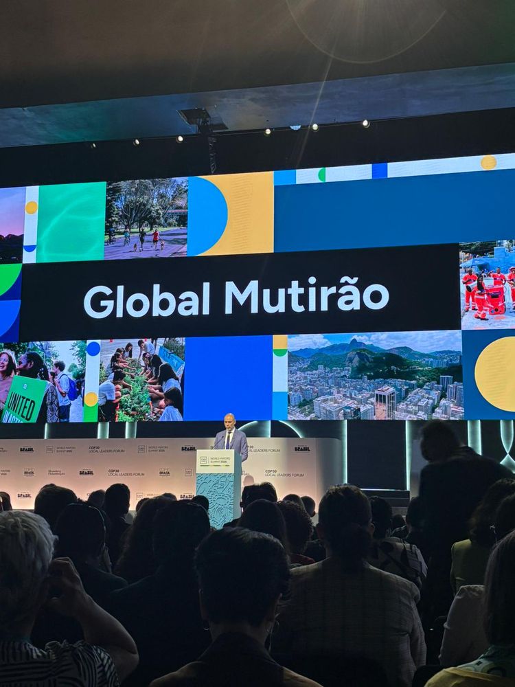 A man speaks at a podium on a stage, facing a large audience. Behind him, a large screen displays the words "Global Mutirão" in white text on a black background, surrounded by a collage of images showing city life, nature, and community activities.
