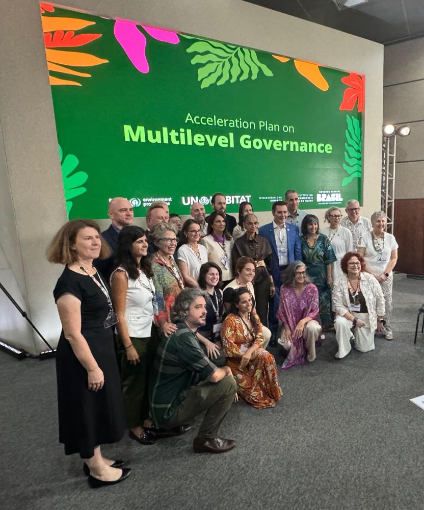 A group of approximately 20-25 people pose for a photograph, standing and kneeling in front of a large green event backdrop. The backdrop reads 'Acceleration Plan on Multilevel Governance' and features logos for UN-Habitat and the Brazilian government.