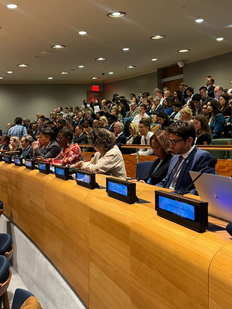 A wide shot of a large, crowded United Nations conference hall filled with a diverse audience. In the foreground, delegates are seated at long wooden desks equipped with microphones and digital placards.