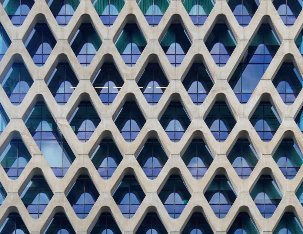 Wabenförmige, hellgraue Betonverkleidung vor der dunkelblauen Glasfassade eines Gebäudes 