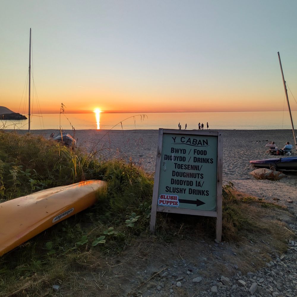 A photo of the sunset at Tresaith beach. There's an advertising board on the grass. People in the background towards the front of the beach look like they're little people stood along the top of board.