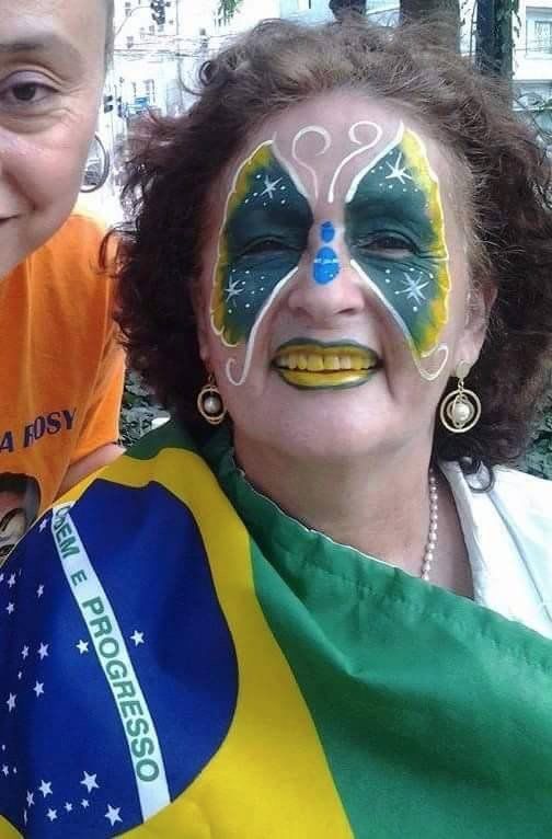 Senhora de idade branca exibindo uma maquiagem facial de borboleta com as cores do Brasil em guache e batom, enrolada em uma bandeira do Brasil