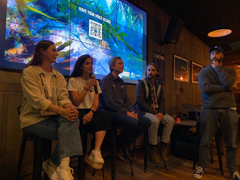 A panel of two women and two men (from left to right) sit on barstools in front of a screen with an underwater photo from the Great Southern Reef and a QR code under the words MAKE YOUR VOICE HEARD. One of the women is holding a microphone, and the panel moderator is standing on the right of the group looking extremely thoughtful. 