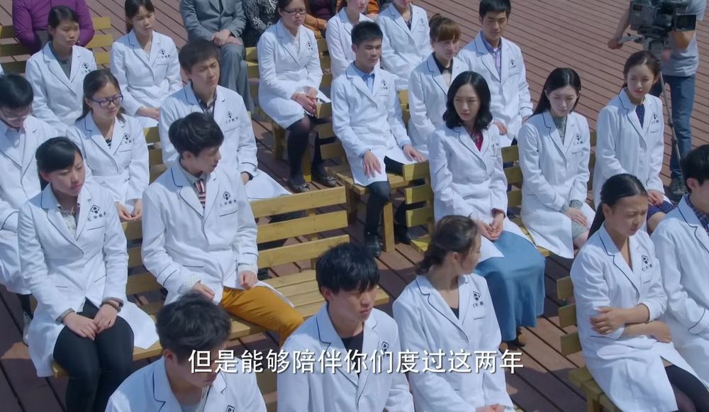 Screenshot from an episode of Grow Up. High shot over rows of medical students sat on benches. There is an empty space by Xie Nanxiang and he has turned to look at it, as has Ye Chunmeng who is in the next row over.