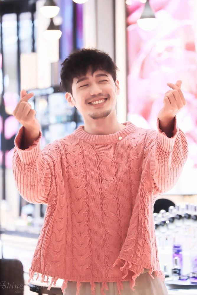 Bai Yu smiling and making finger hearts, wearing a pink knitted jumper
