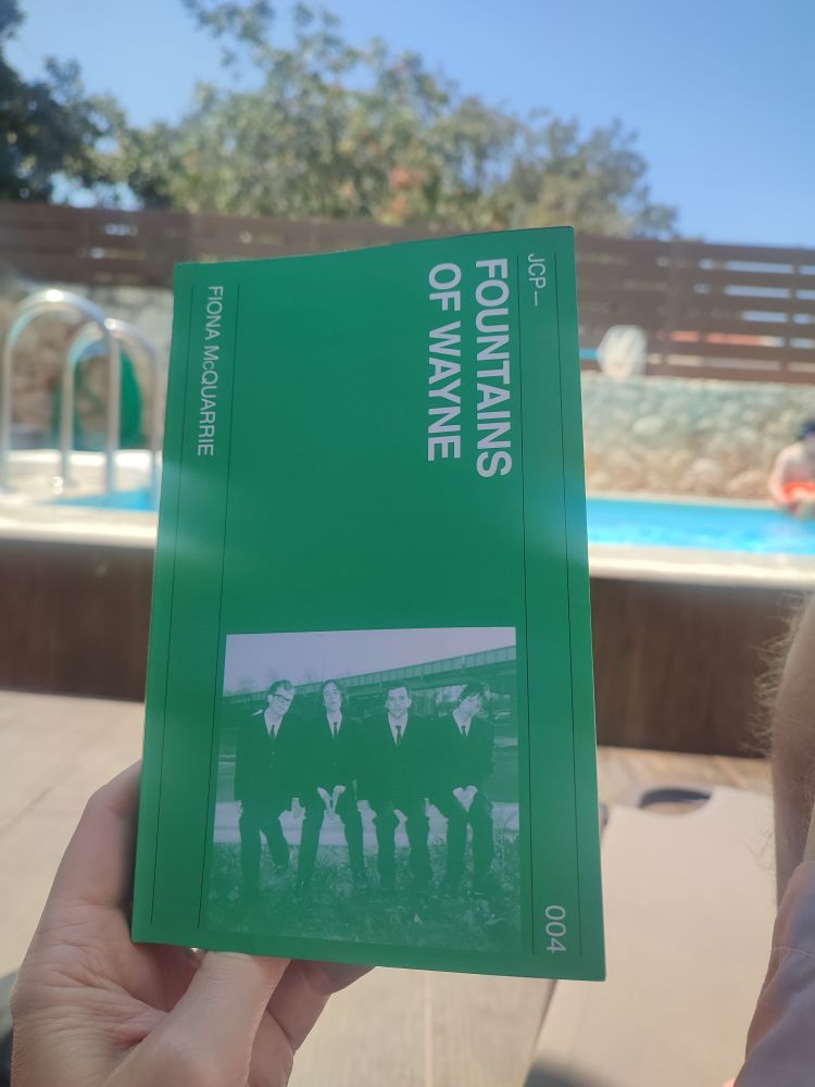 The book Fountains of Wayne by Fiona McQuarrie being held in front of a swimming pool and a blue sky