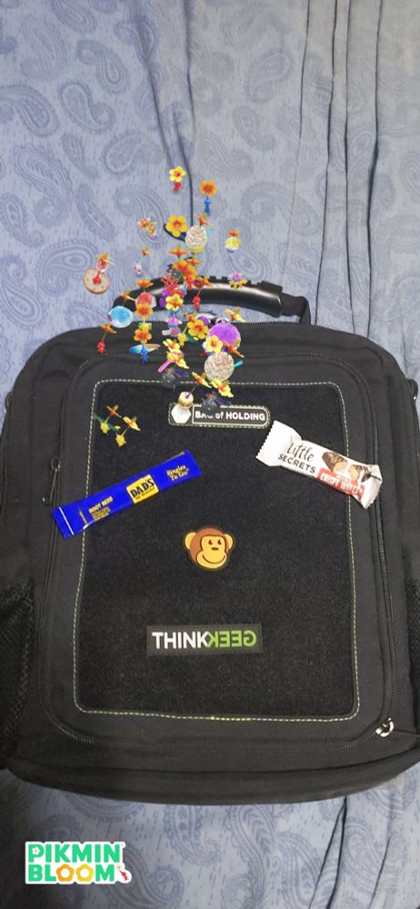 Photo of a ThinkGeek Bag of Holding specifically for carrying electronic devices during conventions. It's all black with the logo and monkey mascot, and a packet of Dad's Root Beer drink mix and a tiny peanut butter wafer candy bar rest on top of it. A squad of Pikmin blooming yellow hibiscus flowers on their heads are milling around at the top.