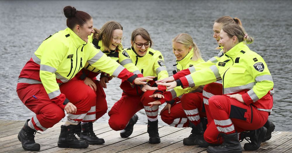 Kuusi ensihoitajaa kyykyssä laiturilla puolipiirissä pitävät käsiään yhdessä keskellä.