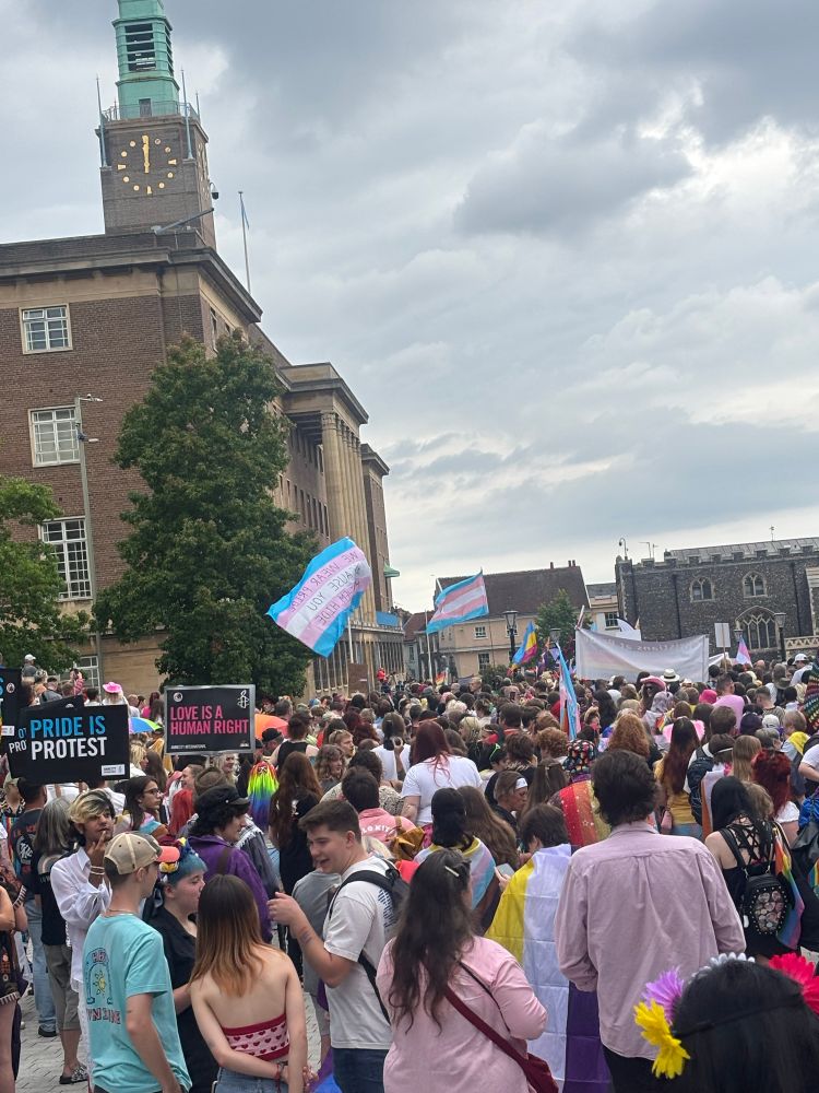 A crowd gathered in front of Norwich City hall for the Pride march. Visible are a placard saying ‘Pride is Protest’, one saying ‘Love is a human right’ and several pride flags. 