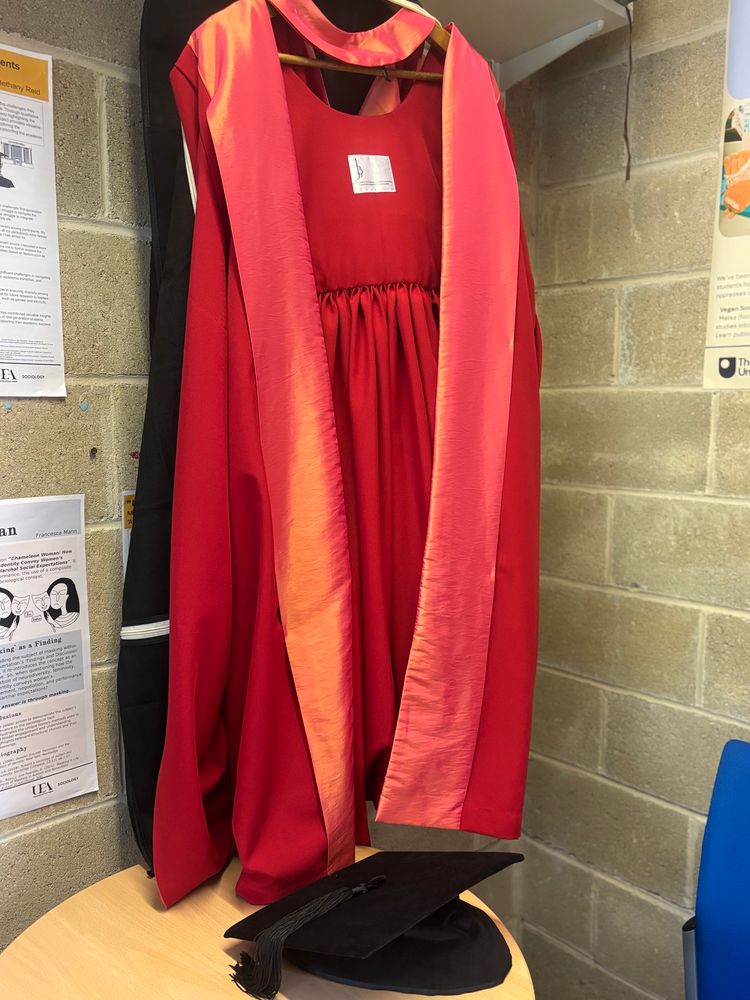 A University of Wales PhD gown and mortarbord hanging in my office. The gown is crimson with linings of red shot with yellow (basically shiny orange. On the table in front of the gown is a black velvet mortarboard. 
