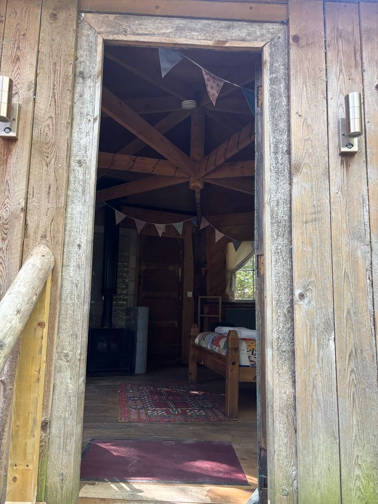 The open door of a treehouse looking inside. There are a coupe of rugs on the floor, the end of a double bed, and bunting hanging from the ceiling. 