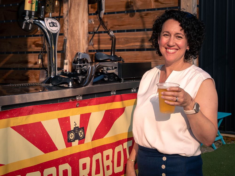 Claudia Chender smiles while holding a beer in front of a Good Robot Brewing Co. tap station at an outdoor event. 
