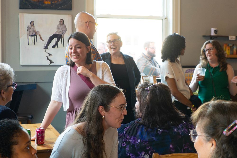 A lively room filled with people chatting and laughing during a community event. A woman in a mauve dress and white cardigan smiles while walking past seated attendees.