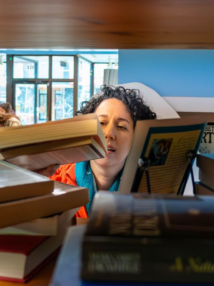 Claudia Chender browses a shelf of books in a brightly lit store. She's partially obscured by stacks of books in the foreground, holding one and reading its back cover.