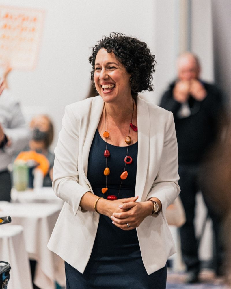 Claudia Chender is smiling and walking into the room having just been announced as Leader of the NSNDP in a lively indoor setting. She is wearing a navy dress, a white blazer, a colorful chunky necklace with red, orange, and yellow beads, and a watch. The background shows blurred people cheering and clapping at tables covered with white tablecloths.