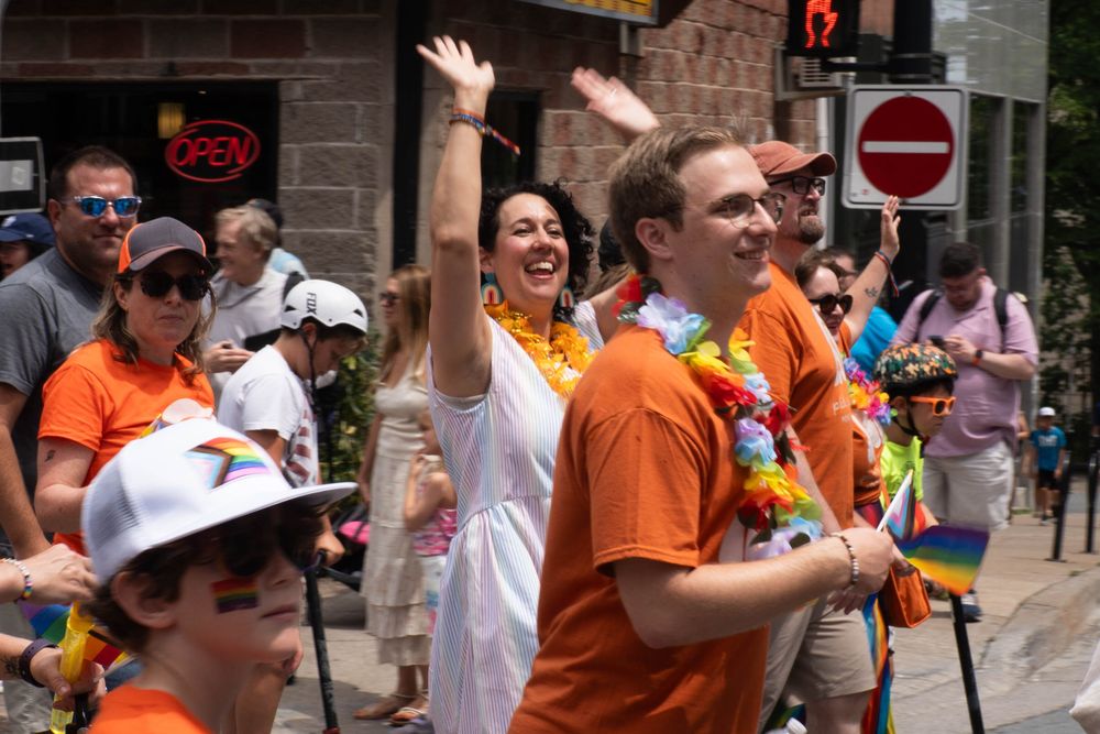 A group of people in orange shirts and rainbow accessories march in a Pride parade. Claudia Chender is in the center raising her arms joyfully while others wave rainbow flags and wear flower garlands.