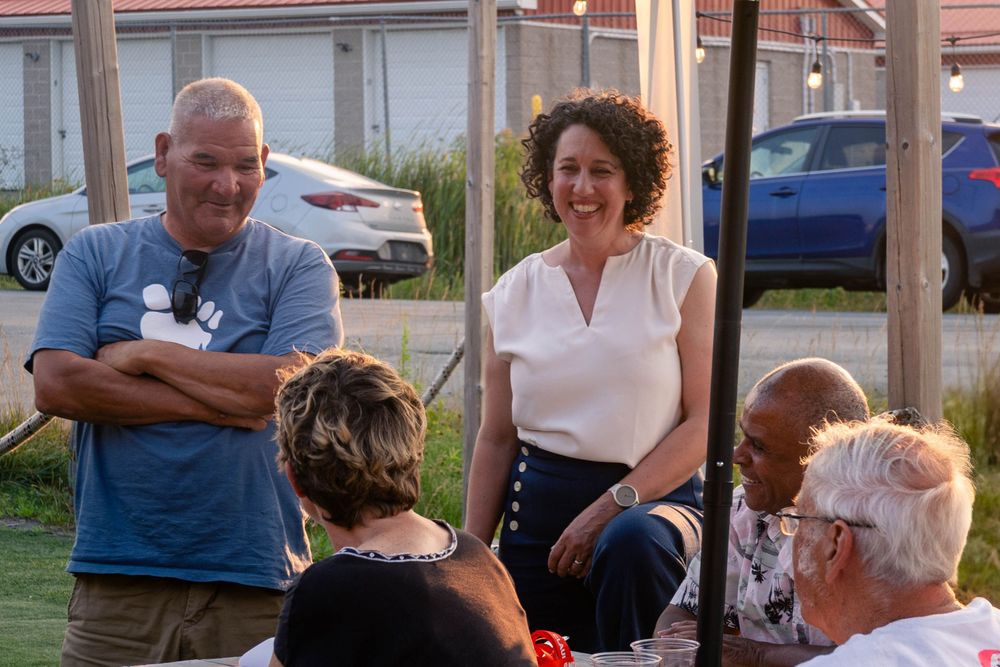 Claudia Chender stands and smiles at a group sitting at a picnic table, while a man in a blue shirt with folded arms stands beside her.