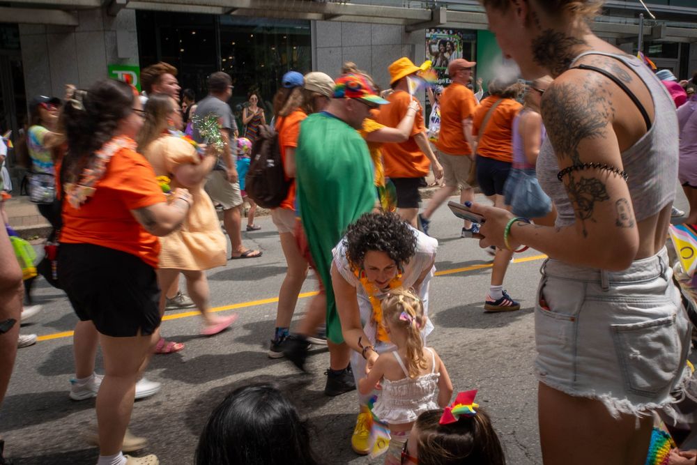 At a Pride parade, Claudia Chender wearing a striped dress bends down to greet a small child dressed in white. Parade participants in bright clothing and rainbow accessories fill the street behind them.