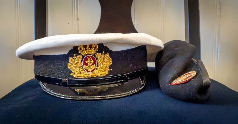 A white-topped naval officer's cap with a gold and red emblem sits on a chair beside a black beret featuring a red-edged insignia. Both hats are displayed neatly indoors, suggesting a formal or ceremonial setting.