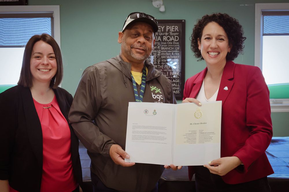 Claudia Chender, Chester Borden, and Kendra Coombes stand together smiling. Chester is in the center wearing a cap and jacket with a “BGC” logo and holds an official certificate. The two women on either side wear blazers, and Claudia is on the right, in a red blazer, also holding the certificate.
