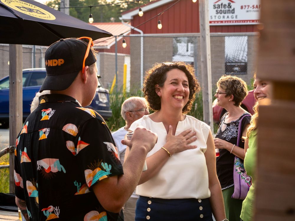 Claudia Chender smiles with her hand on her chest while chatting with two people at an outdoor event. String lights and umbrellas are in the background.