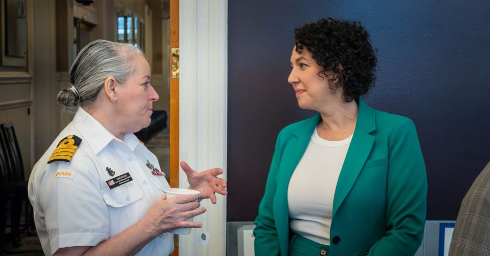 Captain Beth Vallis, Chief of Staff to Commander, Maritime Forces Atlantic in a Royal Canadian Navy uniform is speaking animatedly with Claudia Chender who is wearing a green blazer and white shirt. They are standing indoors in front of a dark blue wall and doorway, engaged in conversation.