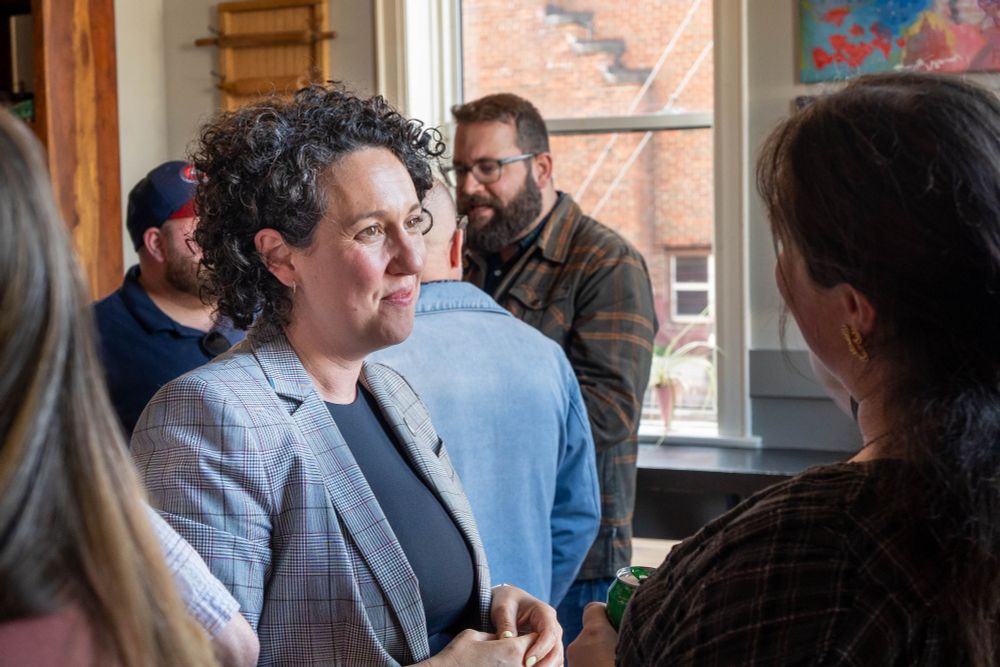 Claudia Chender, in a grey plaid blazer, speaks attentively with another woman in a crowded room. Other attendees are chatting in the background near a bright window.