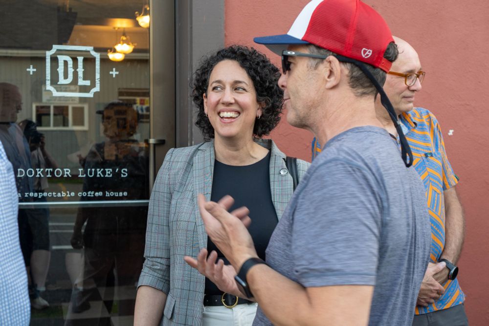 Outside a café called “Doktor Luke’s,” a Claudia Chender in a plaid blazer smiles warmly while talking with a man in a red and white cap. Others chat nearby, enjoying the sunny day.